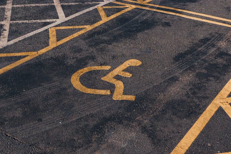A close-up view of a designated accessible parking space on an asphalt surface, featuring a yellow wheelchair symbol painted on the ground. The parking space is marked with yellow lines, with part of a building entrance visible in the background. The surface shows some tire marks and slight wear. This parking area, situated outside a residential or commercial property, is adjacent to a doorway for easy access during a home relocation or moving process. The image highlights the importance of accessible parking arrangements when planning a furniture transportation or home packing and moving service, such as those provided by Man with Van Sydenham Hill, ensuring efficiency and compliance during house removals on College Road SE26.