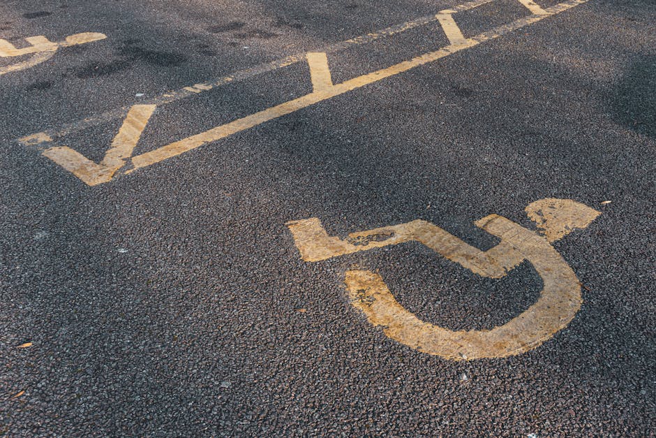 Close-up image of a disabled parking space painted on an asphalt surface, featuring a yellow wheelchair symbol and white dashed lines marking the designated area. The surface appears textured with small gravel particles, and the yellow paint shows signs of wear and fading. This parking space is situated outdoors, likely on a street or in a parking lot adjacent to a residential or commercial property. The image captures the area used for vehicle loading and unloading, which is relevant to house removals and furniture transport services. Man with Van Sydenham Hill, specializing in relocations and home moving logistics, may utilize such parking zones for efficient loading of furniture, boxes, and appliances during packing and moving processes. Clear and accessible parking arrangements like this are essential for smooth operations when executing house removals, facilitating timely and organized furniture transport from properties to moving vehicles.