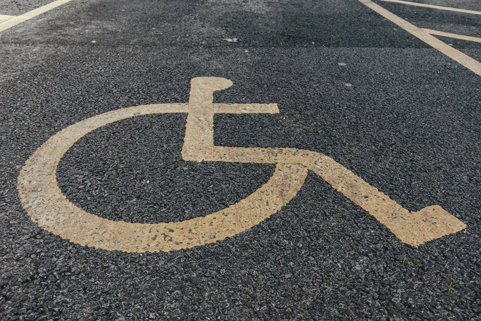 A close-up view of a designated accessible parking space on an asphalt surface, featuring a yellow wheelchair symbol painted on the ground. The parking space is marked with yellow lines, with part of a building entrance visible in the background. The surface shows some tire marks and slight wear. This parking area, situated outside a residential or commercial property, is adjacent to a doorway for easy access during a home relocation or moving process. The image highlights the importance of accessible parking arrangements when planning a furniture transportation or home packing and moving service, such as those provided by Man with Van Sydenham Hill, ensuring efficiency and compliance during house removals on College Road SE26.