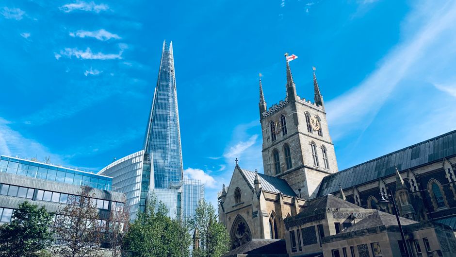 A cityscape featuring the modern, glass-clad Shard skyscraper towering over historic stone buildings, including a church with a tall clock tower and pointed spires, all set against a bright blue sky with scattered clouds. The scene includes reflections on the glass surfaces of nearby office buildings and trees lining the street below, capturing a blend of architectural styles typical of central London. This image relates to house removals and furniture transport, with the existing structures indicating a busy urban environment suitable for interior packing and loading activities by companies such as Man with Van Sydenham Hill, as they carry out home relocation or moving logistics in the area.
