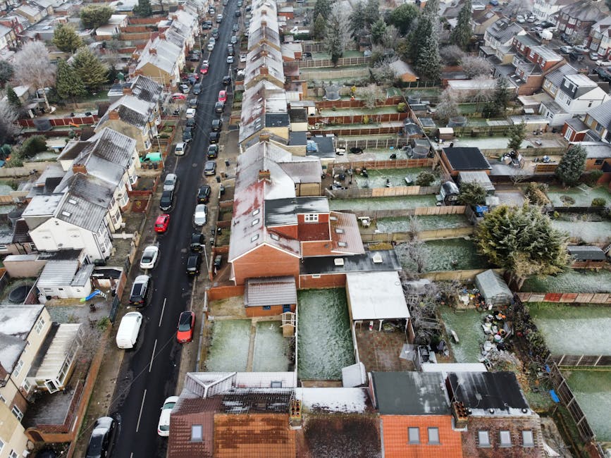 An aerial view of a residential neighbourhood showing a narrow, winding street lined with parked cars on both sides. On either side of the road, there are terraced houses with pitched roofs, some covered with snow or ice. Behind the houses, multiple fenced back gardens are visible, with a variety of outdoor furniture, sheds, and garden features. Some gardens have patio areas, lawns, or vegetable plots, and several contain trees, shrubs, and decorative plants. The image captures a scene typical of home relocation and furniture transport logistics, with the street serving as a potential loading zone for a professional removals team like Man with Van Sydenham Hill, who might be engaged in packing and moving services involving household furniture, boxes, and appliances in preparation for a house move.
