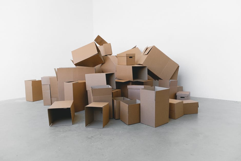 A large collection of various cardboard boxes of different sizes and shapes, some open and some sealed, are stacked and scattered on a light grey concrete floor against a plain white wall. The boxes include cube-shaped, rectangular, and collapsible types, with some placed inside each other or leaning against the wall, indicating a packing or moving process. A few larger boxes are positioned near the wall, while smaller boxes are spread out on the floor, with some stacked on top of others. The environment appears to be an empty room or storage area prepared for home relocation or furniture transport, as associated with house removals services. The scene is well-lit with even lighting, highlighting the materials which primarily consist of plain brown cardboard. This setup suggests the initial stage of packing or loading items for a move, and it reflects the logistical activity involved in furniture transport and packing during relocation, as offered by Man with Van Sydenham Hill.