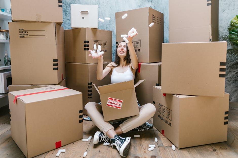 A woman sitting cross-legged on a wooden floor inside a room, surrounded by numerous large cardboard boxes labeled with handling instructions and containing household items. She is smiling and tossing packing foam pellets into the air, with some falling around her. The boxes are stacked against a textured wall, and a small potted plant is visible in the background. The scene depicts the packing and moving process involved in home relocation services, with a focus on packing materials, boxes, and the preparation involved in furniture transport by a removals company such as Man with Van Sydenham Hill.