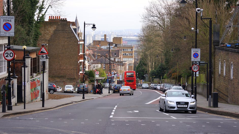 A city street scene showing the entry point of a residential area with several parked cars along both sides of the road and a red double-decker bus approaching from the distance. The street is lined with brick buildings on the left, including a multi-story and a smaller structure, and on the right with trees and a brick wall. There are multiple traffic signs, including controlled zone and speed limit signs, and black bollards along the sidewalk. Street lamps and signage are visible, with some conduct zones and parking restrictions indicated. In the foreground, the road has white dashed lane markings, and the pavement is clear of obstructions. In the background, a slight incline in the road leads towards a cityscape with high-rise buildings under an overcast sky. The scene captures a typical urban environment suitable for highlighting house removals and furniture transport activities, aligned with Lewisham Council van permit rules for Sydenham Hill, as managed by Man with Van Sydenham Hill.