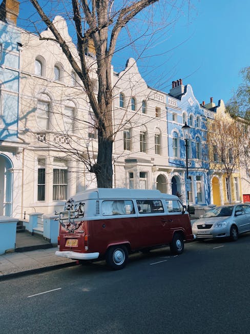 A vintage red and white Volkswagen Type 2 van parked on a residential street in Sydenham Hill, London, with a row of colorful Victorian-style terraced houses in the background. The van is equipped for house removals, featuring a roof rack loaded with metal bicycle frames and a bicycle, and the van's side door is closed. The street has a paved pavement with a narrow curb, and a silver car is parked adjacent to the van. Several bare trees are along the sidewalk, indicating a winter or early spring day, with clear blue sky overhead and sunlight casting shadows on the buildings. This scene illustrates a home relocation process with the van positioned near the property, ready for furniture transport and packing during a house move, demonstrating the logistical aspects of a professional removals service by Man with Van Sydenham Hill, aligned with Lewisham Council van permit rules.