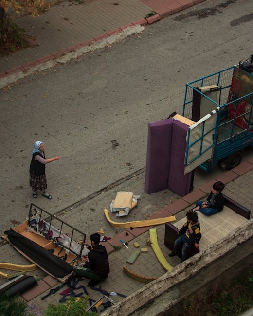 An aerial view capturing a street scene during a home relocation process with visible furniture and packing materials. A blue flatbed truck with an open side gate is parked on the pavement near the property, loaded with large, wrapped items including padded furniture pieces and cardboard boxes, indicating a furniture transport operation. On the sidewalk, three individuals are seated on a bench, observing the activity, while a young girl wearing a hat and dark clothing is standing nearby, possibly waiting or communicating with the movers. Various packing materials such as foam padding, cardboard, and fabric covers are scattered around, along with small tools and straps used for securing items during the move. The environment is a residential street with a curb, red brick edging, and a mixture of pavement and tarmac surface. This scene illustrates the logistical aspects of furniture moving and packing during a house removal carried out by a professional service, such as Man with Van Sydenham Hill, emphasizing the careful handling and organization involved in home relocation.