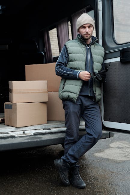A man wearing a beige knit beanie, a light green puffer vest, and a dark jacket with blue and white stripes on the sleeves, standing inside a moving van adjacent to a doorway. He is holding a black st