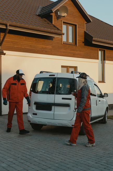 Two removal team members from Man with Van Sydenham Hill are engaged in a home relocation process outside a modern, two-storey house with wooden cladding and large windows. The team, dressed in red un