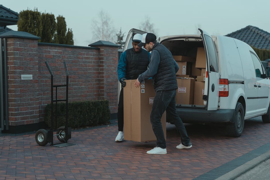 Two removal team members from Man with Van Sydenham Hill are engaged in a home relocation process outside a modern, two-storey house with wooden cladding and large windows. The team, dressed in red un