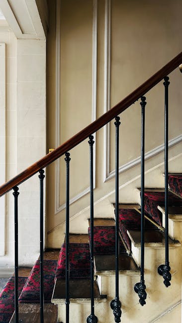 The image shows a staircase inside a Victorian-style home with a wooden handrail and black metal balusters. Red-patterned carpets cover the steps, which are made of wood or stone. The staircase runs alongside a beige wall with decorative molding, and a partially visible door or opening is located at the bottom of the stairs. Natural light illuminates the scene, highlighting the textured surface of the stair treads and the elegant architectural details. The setting appears to be part of a home relocation process, with the staircase ready for moving equipment or furniture transport, as managed by a professional removals service such as Man with Van Sydenham Hill. The environment reflects a typical interior that requires careful navigation and handling of household items during a move.
