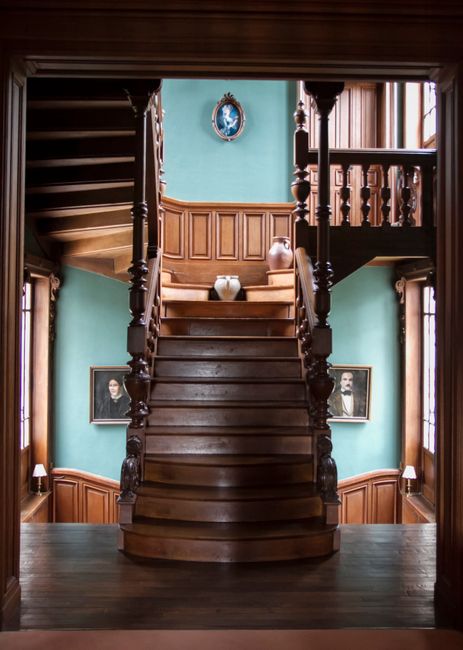 Inside a historic Victorian home, a wide wooden staircase with ornate balustrades and polished steps leads upward from a wooden floor, flanked by teal-painted walls with dark wood paneling. On the staircase, a cardboard box wrapped in plastic sits at the top, surrounded by packing materials and a large ceramic vase. To the sides of the staircase, small tables hold decorative lamps, and two framed black-and-white portraits are hung on the walls, indicating a focus on home decor and preservation. Natural light filters through large windows on the right, illuminating the interior, which features detailed woodwork and classical furnishings. This scene captures the interior of a property undergoing a home relocation or packing and moving process, with visible elements suitable for professional removals services like those provided by Man with Van Sydenham Hill, emphasizing careful handling of furniture and fragile items during a residence move.