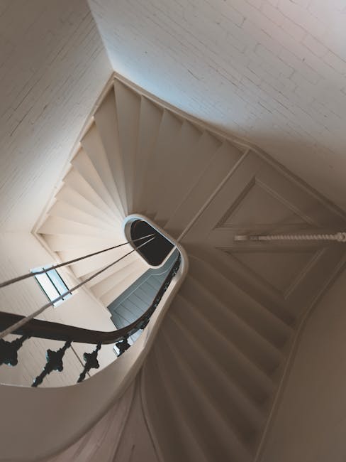 The image shows a staircase inside a Victorian-style home with a wooden handrail and black metal balusters. Red-patterned carpets cover the steps, which are made of wood or stone. The staircase runs alongside a beige wall with decorative molding, and a partially visible door or opening is located at the bottom of the stairs. Natural light illuminates the scene, highlighting the textured surface of the stair treads and the elegant architectural details. The setting appears to be part of a home relocation process, with the staircase ready for moving equipment or furniture transport, as managed by a professional removals service such as Man with Van Sydenham Hill. The environment reflects a typical interior that requires careful navigation and handling of household items during a move.