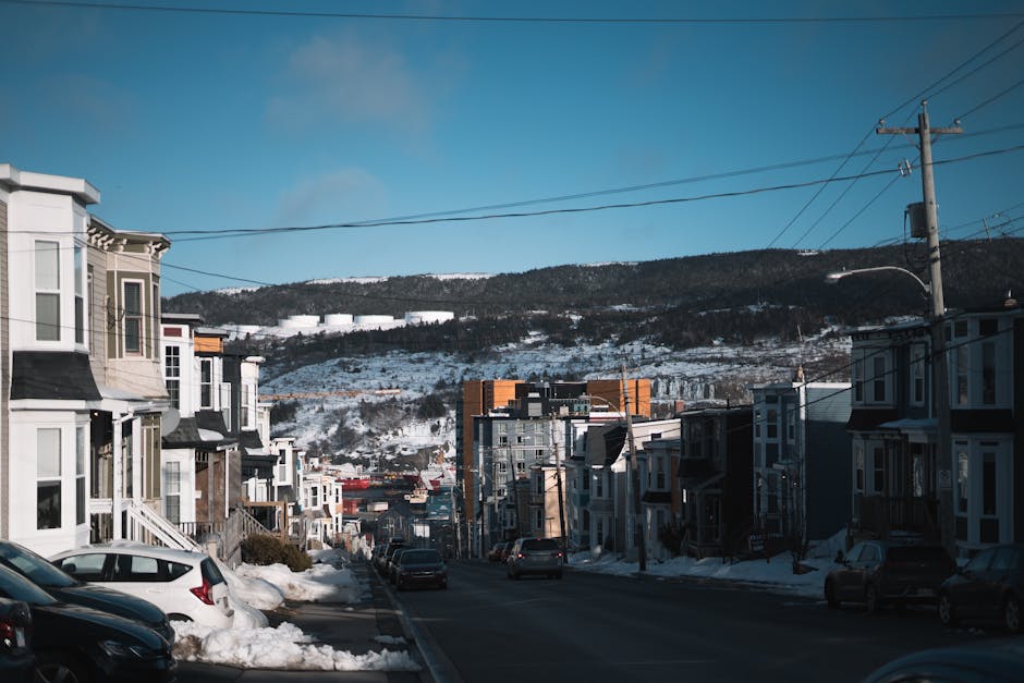 Photograph of a residential street in Sydenham Hill during winter, with snow covering the pavement and surrounding landscaped areas. On the left side, there are multi-storey terraced houses with bay windows, some with white or beige exterior finishes. Parked cars, including a white hatchback and a dark-colored vehicle, line both sides of the street. The road appears clear for movement, with no visible pedestrians or moving vehicles. Overhead, several power lines run parallel to the street, supported by tall wooden utility poles. In the background, a hillside with snow-covered trees and some larger modern buildings or industrial structures at the top is visible against a clear blue sky. This scene illustrates a typical home relocation environment, emphasizing urban setting and transportation aspects relevant to house removals and furniture transport, with clear natural lighting that highlights the winter scene.