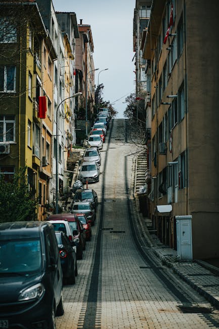 Photograph of a residential street in Sydenham Hill during winter, with snow covering the pavement and surrounding landscaped areas. On the left side, there are multi-storey terraced houses with bay windows, some with white or beige exterior finishes. Parked cars, including a white hatchback and a dark-colored vehicle, line both sides of the street. The road appears clear for movement, with no visible pedestrians or moving vehicles. Overhead, several power lines run parallel to the street, supported by tall wooden utility poles. In the background, a hillside with snow-covered trees and some larger modern buildings or industrial structures at the top is visible against a clear blue sky. This scene illustrates a typical home relocation environment, emphasizing urban setting and transportation aspects relevant to house removals and furniture transport, with clear natural lighting that highlights the winter scene.